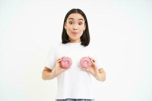 glimlachen gelukkig Aziatisch vrouw lachend, Holding twee geglazuurd donuts over- borst, staand over- wit achtergrond foto