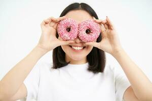 beeld van grappig jong vrouw, maakt bril met twee roze geglazuurd donuts, op zoek door donut gaten en lachend, geïsoleerd Aan wit achtergrond foto