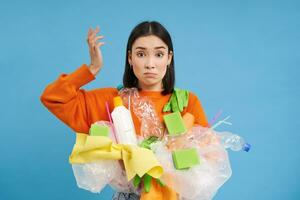 verward Koreaans vrouw probeert naar Gaan groente, recycle plastic huishouden artikelen, Holding flessen en handschoenen, doet recyclen, blauw achtergrond foto