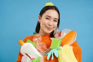 portret van enthousiast Koreaans vrouw, Holding leeg plastic flessen, sorteren afval, glimlachen en op zoek Vrolijk, geïsoleerd Aan blauw achtergrond foto