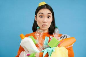 portret van Aziatisch vrouw met gefascineerd gezicht, houdt onzin voor recyclen, leeg plastic flessen en afval, staart verward, blauw achtergrond foto