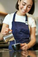 verticaal schot van brunette Aziatisch meisje barista, vervelend schort, maken koffie, bereiden bestellen in cafe, gieten melk in kop voor cappuccino latte kunst foto