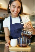 verticaal schot van glimlachen schattig barista meisje, gieten filter koffie, maken bestellen voor cafe cliënt, vervelend blauw schort foto