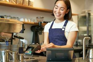 glimlachen Aziatisch meisje barista, geven cliënt pos terminal, helpen betalen met credit kaart, staand Bij teller in koffie winkel, werken in cafe foto