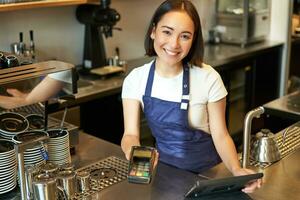 portret van mooi Aziatisch meisje barista, koffie winkel personeel geeft pos terminal, helpt betalen contactloos met credit kaart, staand Bij teller in cafe foto