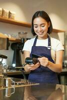 portret van glimlachen Aziatisch barista, koffie winkel werknemer gebruik makend van pos terminal en credit kaart, helpt cliënt betalen contactloos in cafe foto