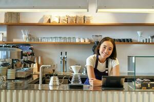 gelukkig glimlachen Aziatisch barista, meisje achter balie, werken met pos terminal en brouwen filter uitrusting, maken koffie in cafe foto