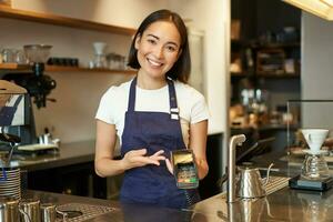 portret van Aziatisch barista meisje Bij balie, tonen kaart machine naar cliënt wie wil naar betalen contactloos, nemen bestellen, staand in cafe foto