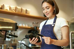 schattig Aziatisch meisje, barista in cafe verwerken contactloos betaling, insert credit kaart in pos terminal, nemen bestellen in koffie winkel foto