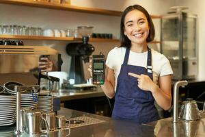 glimlachen Aziatisch barista meisje, draagt schort, shows credit kaart machine voor verwerken betaling, stel voor naar betalen contactloos, staand in koffie winkel foto