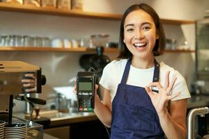 glimlachen Aziatisch barista meisje, draagt schort, shows credit kaart machine voor verwerken betaling, stel voor naar betalen contactloos, staand in koffie winkel foto