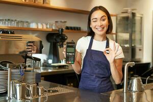 glimlachen Aziatisch barista meisje, draagt schort, shows credit kaart machine voor verwerken betaling, stel voor naar betalen contactloos, staand in koffie winkel foto