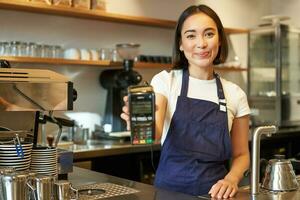 glimlachen Aziatisch barista, koffie winkel personeel geeft u credit kaart machine, verwerken betaling met pos terminal, werken in cafe foto