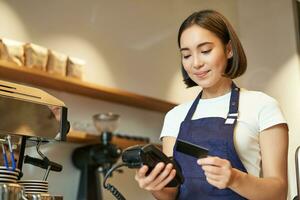 schattig Aziatisch meisje, barista in cafe verwerken contactloos betaling, insert credit kaart in pos terminal, nemen bestellen in koffie winkel foto