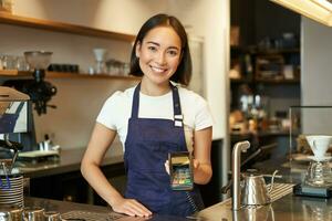 portret van Aziatisch barista meisje Bij balie, tonen kaart machine naar cliënt wie wil naar betalen contactloos, nemen bestellen, staand in cafe foto