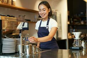 glimlachen meisje barista in cafe, voorbereidingen treffen cappuccino in koffie machine, stomen melk, vervelend uniform schort foto