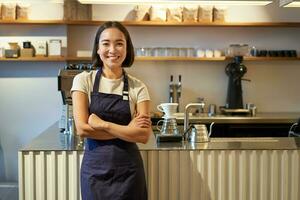 portret van glimlachen Aziatisch vrouw barista, vervelend schort, staand in de buurt teller met koffie, werken in cafe foto