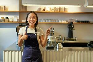 gelukkig jong Aziatisch barista, vrouw personeel arbeider in schort, tonen duimen omhoog en richten Bij kopiëren ruimte, demonstreren cafe advertentie foto