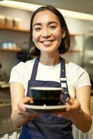 verticaal schot van vriendelijk Aziatisch meisje lachend, portie koffie, barista geven u kop van koffie, bereid drinken voor cliënt in cafe foto