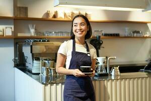 portret van Aziatisch glimlachen vrouw barista, cafe serveerster Holding kop van koffie, portie klanten, nemen bestellen naar de tablet, staand in de buurt cafe teller foto