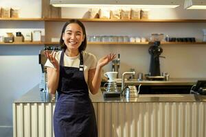 enthousiast jong Aziatisch vrouw, barista in schort, op zoek versteld staan, klap handen en lachend, staand in koffie winkel, werken in cafe foto