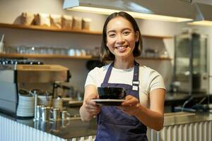 portret van glimlachen Aziatisch vrouw houdt kop van koffie, bereiden drankjes voor klanten in cafe, werken en portie drankjes, vervelend uniform schort foto