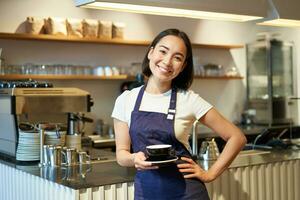 portret van glimlachen Aziatisch vrouw barista, maken koffie, Holding kop van thee en nemen het naar cafe cliënt, vervelend schort, staand in de buurt teller foto
