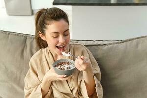 portret van prachtig brunette vrouw zit Aan bank, hebben haar ontbijt Bij huis, glimlachen terwijl aan het eten granen, Holding kom en lepel en lachend foto