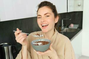 gelukkig ochtenden. prachtig jong vrouw aan het eten granen met melk, staand in keuken met ontbijt schaal, genieten van begin van de dag, glimlachen foto