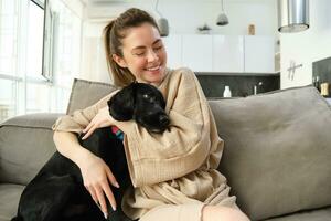 geluk en levensstijl concept. aanbiddelijk hond besteedt tijd met haar baasje, jong vrouw knuffelen haar puppy Aan sofa Bij huis foto