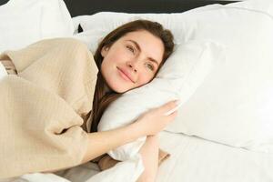 prachtig vrouw resting in bed vervelend pyjama, knuffelen haar hoofdkussen en glimlachen foto