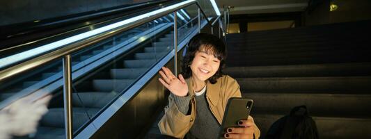 portret van Koreaans meisje zit Aan trap en golven hand- Bij mobiel telefoon, video chats Aan smartphone, groeten vriend Aan sociaal media foto