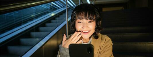 Koreaans meisje looks verrast en gelukkig Bij haar telefoon scherm, glimlacht versteld staan, leest mooi zo nieuws, zit Aan trappenhuis in de buurt roltrap in winkelcentrum, luistert muziek- in hoofdtelefoons foto