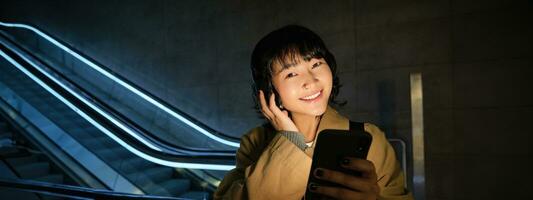 portret van mooi Koreaans meisje in koptelefoon, staat in de buurt roltrap, reizen, pendelt huis Aan openbaar vervoer, houdt smartphone, poses met mobiel telefoon foto