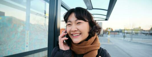 dichtbij omhoog van schattig Koreaans vrouw, maken een telefoon telefoongesprek, pratend en glimlachen Aan telefoon, staand in winter jasje Aan bus hou op, aan het wachten voor haar vervoer naar aankomen foto