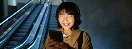 lachend schattig Koreaans meisje Aan roltrap, gaan naar beneden naar buis, ondergronds of metro, gebruik makend van mobiel telefoon, aan het kijken iets grappig Aan smartphone app foto