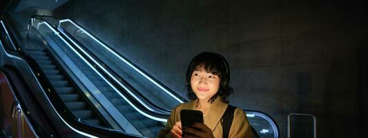 glimlachen Koreaans meisje in koptelefoon, luistert muziek, toepassingen mobiel telefoon in buis, staat in de buurt roltrap in donker, looks vrolijk foto