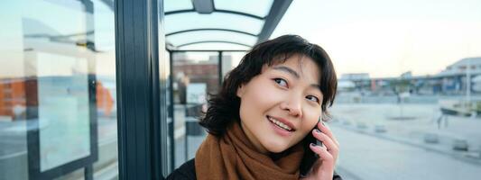 dichtbij omhoog van schattig Koreaans vrouw, maken een telefoon telefoongesprek, pratend en glimlachen Aan telefoon, staand in winter jasje Aan bus hou op, aan het wachten voor haar vervoer naar aankomen foto