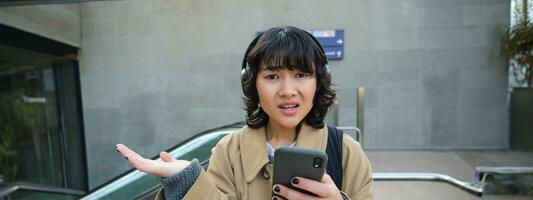 dichtbij omhoog portret van verbaasd, ingewikkeld Koreaans meisje in koptelefoon, fronst en looks onzeker, staat met smartphone en looks Bij camera terwijl haalt zijn schouders op Aan straat foto