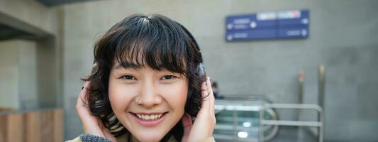 dichtbij omhoog portret van elegant Koreaans meisje staand Aan straat, luisteren muziek- in hoofdtelefoons en lachend, poseren in stad centrum foto
