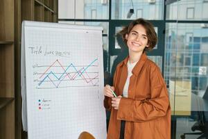 portret van geslaagd zakenvrouw, modern manager in kantoor, staand in de buurt bord met diagram, geven presentatie, geven toespraak in conferentie kamer foto