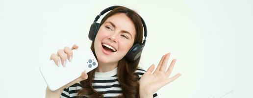 technologie en sociaal media. portret van gelukkig jong vrouw het zingen karaoke, spelen muziek- app Aan smartphone, vervelend draadloze koptelefoon, staand over- wit achtergrond foto