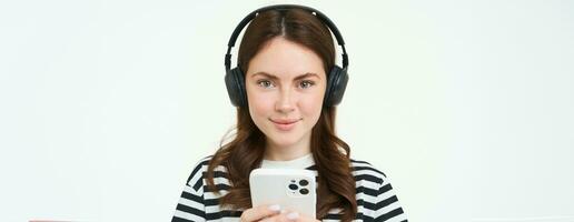beeld van brunette jong vrouw, lachend, luisteren naar muziek- in koptelefoon, aan het kijken videos Aan mobiel telefoon app, Holding smartphone, staand over- wit achtergrond foto