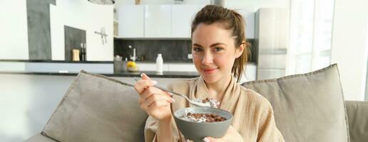 dichtbij omhoog portret van schattig vrouw Aan bank, aan het eten ontbijt, Holding heerlijk chocola granen met melk in een schaal, hebben gepast ochtend- maaltijd foto