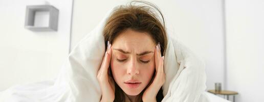 dichtbij omhoog van vrouw gevoel onwel Bij huis, aan het liegen in bed onder wit dekens en fronsend, aanraken hoofd, heeft hoofdpijn, migraine, herstellen van covid foto