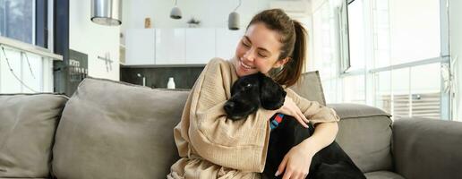 dieren en levensstijl concept. gelukkig jong vrouw in badjas, knuffels haar hond Aan bank, knuffelen puppy en glimlachen foto