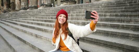 elegant jong meisje in rood hoed, duurt foto's Aan smartphone camera, maakt selfie net zo ze zit Aan trap in de buurt museum, poseren voor foto met app filter