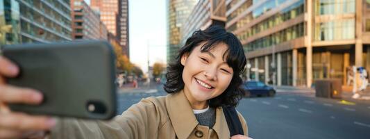 portret van jong Aziatisch vrouw nemen selfie in voorkant van gebouw in stad centrum, toerist duurt foto's terwijl bezienswaardigheden bekijken, glimlachen Bij smartphone camera foto