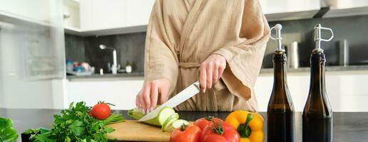 gezond levensstijl. jong vrouw in badjas voorbereidingen treffen voedsel, hakken groenten, Koken avondeten Aan keuken balie, staand over- wit achtergrond foto