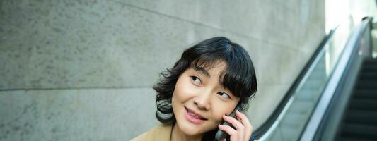 dichtbij omhoog portret van glimlachen brunette meisje, Koreaans vrouw gaat naar beneden roltrap, maakt telefoon telefoongesprek, praat naar iemand Aan telefoon foto